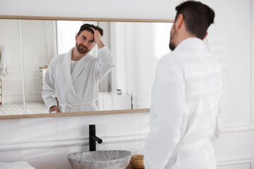 Fototapeta premium Handsome man wearing bathrobe and looking at himself in mirror indoors