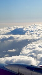 Airplane wing and engine section seen from a window, flying above a vast ocean of fluffy white cumulus clouds with clear blue sky on a travel and holiday journey