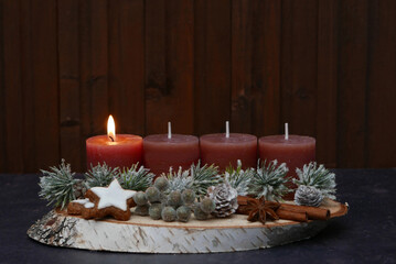 Erster Advent: Längliches Adventsgesteck mit braunen Kerzen, Zimtsternen, Zapfen, Beeren, Zimtstange und Tannenzweigen vor einer braunen Holzwand mit Textfreiraum.