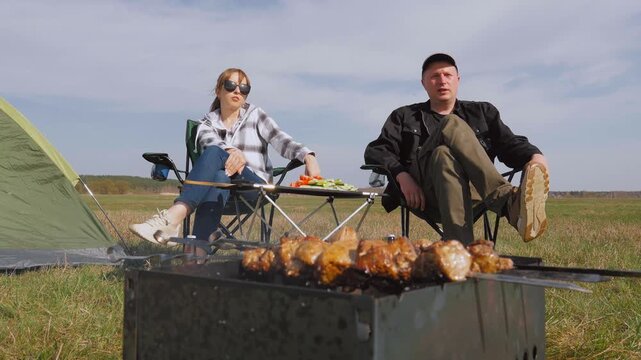 Young couple talking and enjoying themselves while sitting on folding chairs near a tent and grilling delicious shish kebab meat on skewers over a brazier during a picnic in a field