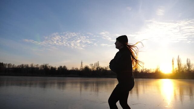 Female skater shod in figure skates sliding on ice surface during beautiful sunset. Sportswoman skating on frozen river. Silhouette of woman training at lake. Shining sun reflecting on surface of ice