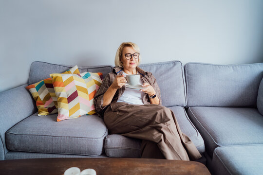 Middle aged smiling woman sitting on sofa and looking away while drinking coffee or tea. Mature senior woman relaxing after housekeeping, home cleaning. Portrait of relaxed female resting at home.