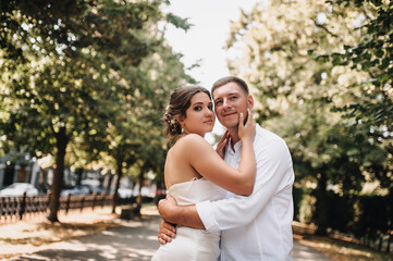 Naklejka premium Wedding photo of happy smiling newlyweds, stylish groom and beautiful young brunette bride, hugging outdoors on the street in the city, in the park.