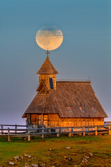 Wooden alpine chapel "Marija snezna" with a cross on the roof, illuminated by warm light, with a super full moon perfectly aligned above the steeple at dusk.