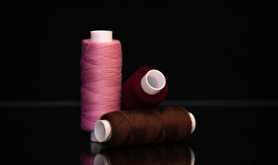 Three sewing thread spools in burgundy, pink and brown colors on a black background. Concept of tailoring, handmade craft, textile materials and fashion design accessories.