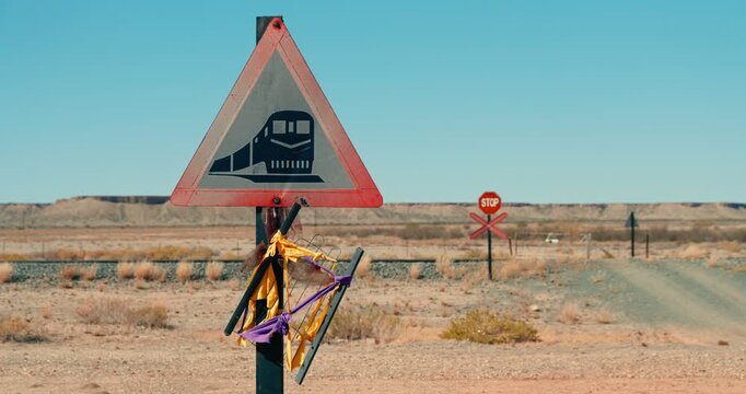 Railway crossing sign and rusty stop sign stand beside gravel road and tracks in Namibia&rsquo;s red desert under blue sky, evoking a stark, remote, dystopian atmosphere.