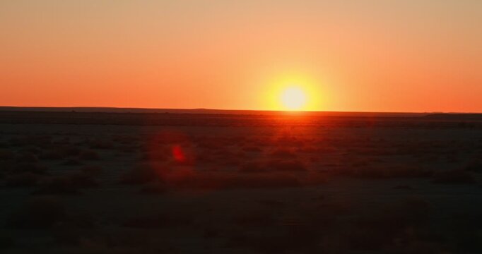 Shot from a moving car of an orange sunset right at the horizon, with the Namibian desert in the foreground, capturing warm fading light across the open landscape.
