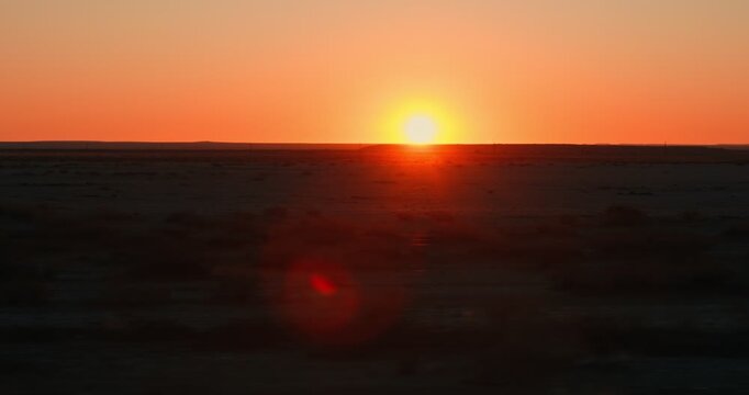 Shot from a moving car of an orange sunset right at the horizon, with the Namibian desert in the foreground, capturing warm fading light across the open landscape.