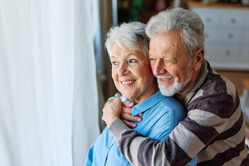 Portrait of a happy senior couple embracing hugging and bonding looking through the window at home