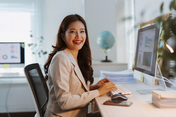 Young Asian businesswoman smiling, looking at camera while typing on a computer keyboard, analyzing data and reports