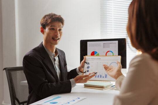 Asian man presenting loan comparison calculator data charts on a clipboard to a woman, discussing statistics in an office meeting