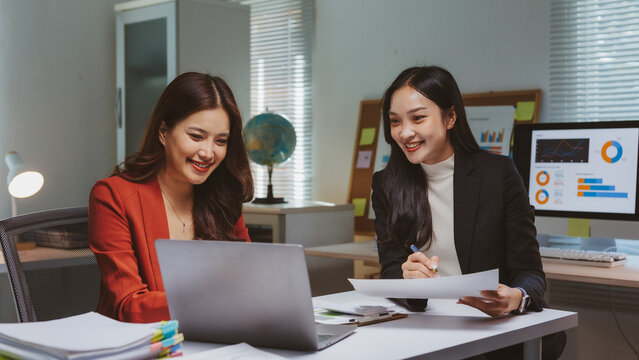 Two smiling Asian businesswomen engaging in a joint discussion, working together on a laptop and analyzing business data in the office