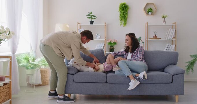 Family parents and daughter play at home. Mom and dad tickle their giggling child girl on sofa, filling the bright room with warmth, smiles, and happiness. Playful family bonding and fun concept.