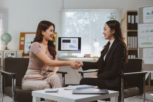 Two smiling Asian businesswomen engaging in a handshake, symbolizing partnership, agreement, and cooperation in a modern office