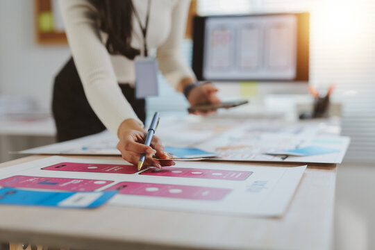 Designer's hand reviewing and annotating printed wireframes for a new mobile application user interface layout, holding a smartphone