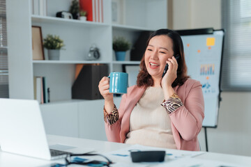 Happy businesswoman talking on smartphone and holding a mug, taking a coffee break in a modern office