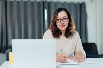Young Asian woman focused on laptop, taking notes in notebook while working or studying remotely from her home office desk