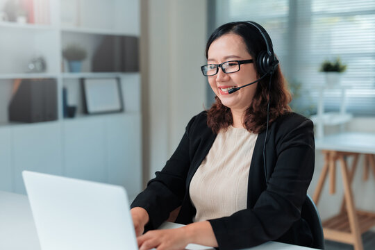 Asian woman operator wearing a headset with microphone, smiling while typing on laptop during remote work or online meeting