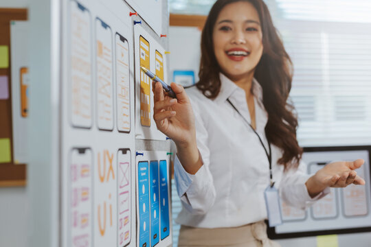 Young Asian woman user experience designer explaining mobile application development mockups and wireframes pinned on a board