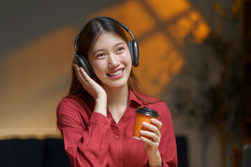 Young Asian woman smiling, listening to music on headphones, and holding a takeaway coffee cup, happy and relaxing