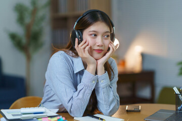 Young Asian woman wearing headphones, listening to music and daydreaming while taking a break from studying at her desk