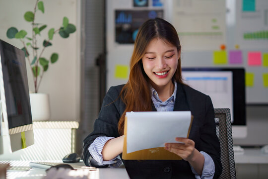 Young professional Asian woman joyfully working, analyzing report papers at her modern office desk, feeling happy and productive - Powered by Adobe