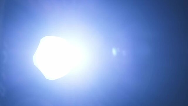 Hand holding a powerful flashlight, pointing its bright beam directly into the camera lens, creating an intense flare and a sense of being found or investigated.