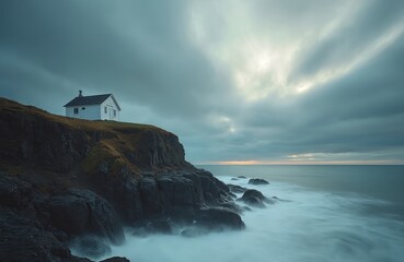 Small white house sits on tall cliff. Rough sea waves crash on dark coastal rocks, creating soft misty effect. Overcast sky with light breaks above vast ocean horizon, showing serene, untamed nature