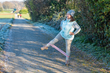 mature plus size woman performing exercise with pink resistance band outdoors in park on cold sunny day strengthening legs and glutes healthy lifestyle and fitness workout, daily outdoor exercises