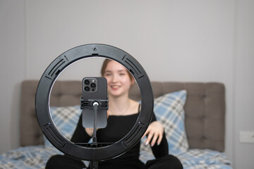 Young girl vlogger recording video with smartphone using ring light while sitting on bed at home, creating engaging content for social media platforms. Focus on the ring lamp