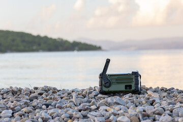 Fototapeta premium Rugged Green Radio Resting On Pebble Shore By Calm Sea, Outdoor Communication Gear