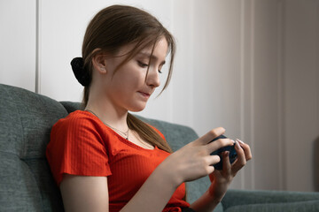 Worried young girl is reading upsetting news on her smartphone while sitting on a gray sofa in her living room, experiencing a moment of concern and uncertainty
