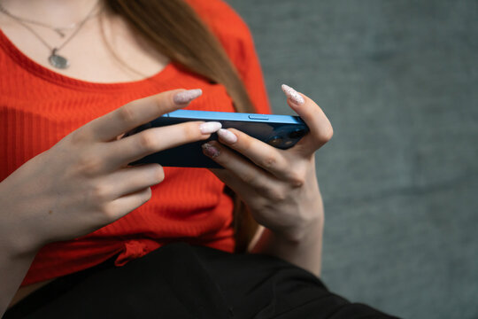 Close-up of female hands holding blue smartphone and playing mobile game, wearing red casual clothes and sitting comfortably, with a blurred gray wall background - Powered by Adobe