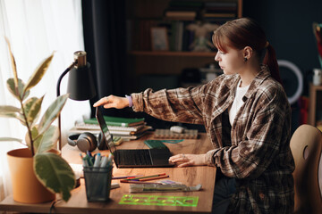 Caucasian teenage girl sitting at desk using laptop, reaching to open computer while studying in room, surrounded by school supplies and books, focused on academic activities