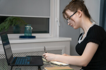 Focused young girl wearing glasses is taking notes in notebook while studying or working remotely using laptop at glass table in cozy home environment