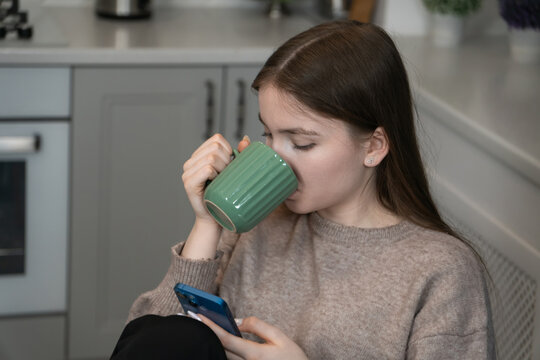 Comfortable teenage girl sipping a hot beverage while scrolling through her smartphone in a modern kitchen, enjoying a perfect blend of relaxation and digital connection during a peaceful morning