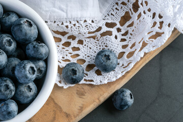 Flat lay still life of fresh blueberries in a white bowl on a wooden board with a lace napkin. Rustic, natural food styling with copy space on dark background.
