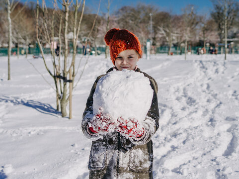 Smiling girl in a winter duffel coat, bobble hat and gloves standing in a park carrying a heavy snowball to make a snowman, Ireland