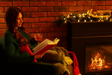 Woman with tea reading book near fireplace in festive decorated room