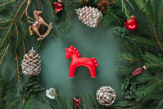 Overhead view of a red horse among festive Christmas baubles, ornaments, pinecones and fir branches on a green background