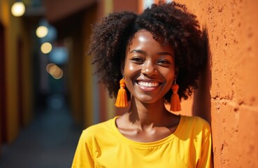 Fototapeta premium Black woman smiles near orange wall. She wears yellow shirt and big earrings. Curly afro hair. Joyful person on a sunny day. Portrait photo.