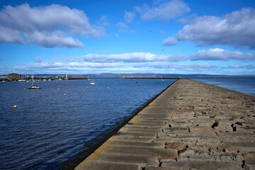 Calm scene along Granton Harbour's stone breakwater, boats dot the distance under blue skies.