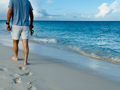 Rear view of a barefoot man walking along a tropical sandy beach carrying his flip flops, Eagle Beach, Aruba