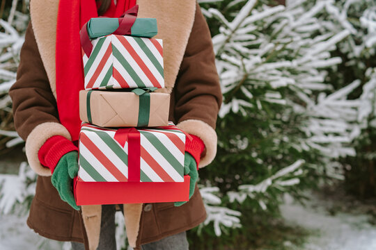 Close-up of a woman in a warm coat and gloves standing by snow covered Christmas fir trees holding a stack of wrapped Christmas gifts