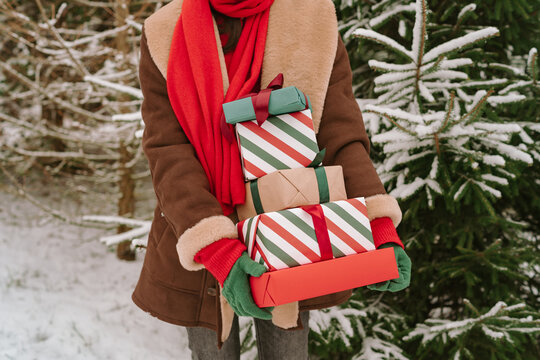 Close-up of a woman in a warm coat and gloves standing by snow covered Christmas fir trees holding a stack of wrapped Christmas gifts