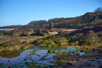 Waves recede on a rugged shore at Seacliff Beach in East Lothian, Scotland, revealing tidal pools and mossy rocks beneath a bright sky.
