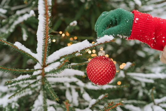 Close-up of a woman in a winter coat and gloves standing outdoors decorating a snow covered Christmas fir tree with a Christmas bauble