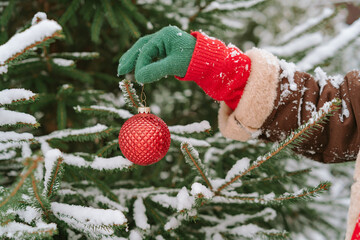 Close-up of a woman in a winter coat and gloves standing outdoors decorating a snow covered christmas fir tree with a christmas bauble