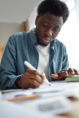 Young Black man analyzing invoices and bills at desk, holding pen and using calculator, focusing on...