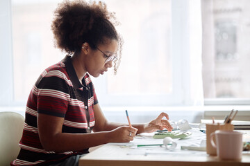 Young Black woman analyzing invoices and bills at desk, holding pencil and reviewing financial documents, focused on paperwork with calculator and office supplies nearby